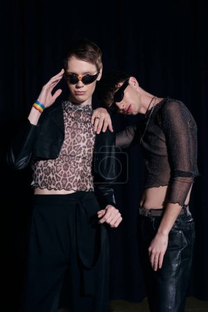 Trendy and young gay friends in sunglasses and party outfits posing and looking at camera during lgbt pride month celebration isolated on black