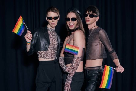 Fashionable and positive homosexual friends in party outfits and sunglasses posing with lgbtq flags during pride month celebration on black background 