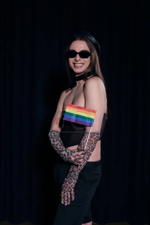 Unbekümmerter und langhaariger schwuler Mann mit Sonnenbrille, sexy Oberteil und Handschuhen, der die lgbt-Fahne während der Pride Community Monatsparty hält, isoliert auf schwarz