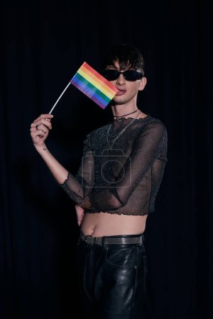 Joven homosexual de moda con gafas de sol y brillante bandera lgbt morder superior y posando durante la celebración del mes de la comunidad orgullo aislado en negro 