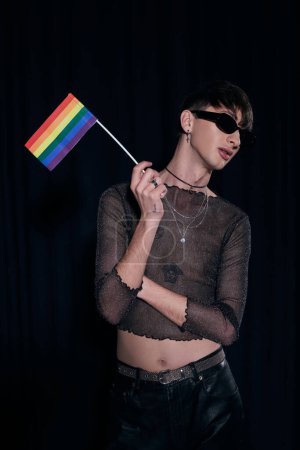 Young and fashionable queer person in sunglasses and shiny top holding lgbt flag while posing during pride community month celebration isolated on black 