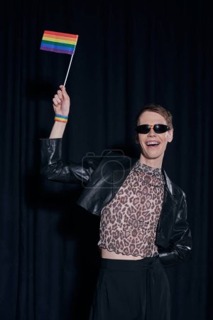 Cheerful and fashionable nonbinary person in leather jacket and top with animal print holding lgbt flag during pride month celebration on black background 