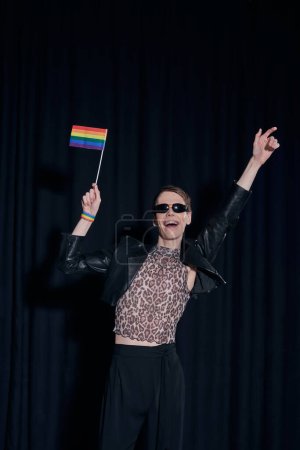 Excited nonbinary person in stylish outfit and sunglasses holding lgbt flag during party and celebration of pride month on black background 