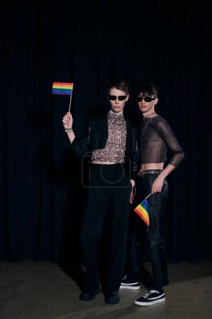 Full length of fashionable gay friends in sunglasses and party outfits holding rainbow lgbt flags during pride community month celebration on black background 