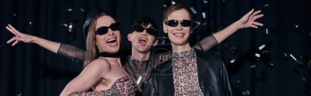 Group of cheerful and trendy homosexual friends in party outfits and sunglasses standing under falling confetti during lgbt month celebration on black background, banner 