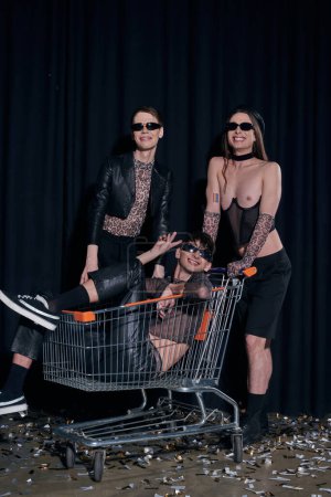 Smiling group of homosexual friends in stylish party outfits having fun with shopping cart during lgbt month celebration on black background 