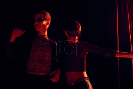 Modische nichtbinäre Freunde in Party-Outfits und Sonnenbrille tanzen zusammen und feiern lgbt Stolz Monat auf schwarzem Hintergrund mit roter Beleuchtung 