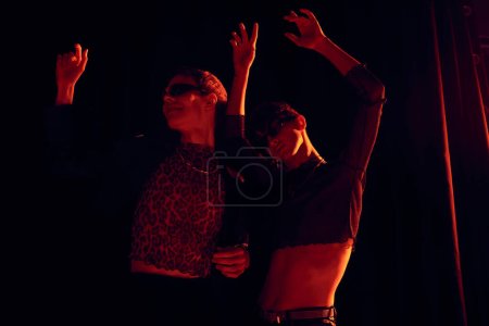 Amigos homosexuales de moda en gafas de sol y trajes de fiesta bailando mientras celebran lgbt comunidad orgullo mes sobre fondo negro con iluminación roja 