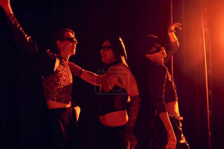 Smiling and fashionable homosexual people in sunglasses dancing during party and lgbt pride month celebration on black background with red lighting 