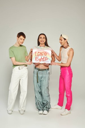 Smiling and young nonbinary friends holding placard with lgbtq rights lettering and standing together while celebrating pride community month on grey background 