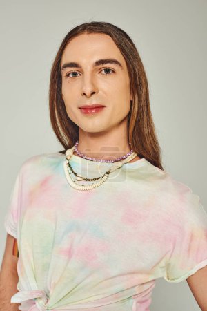 Portrait of young and long haired homosexual man in tie dye t-shirt looking at camera isolated on grey, happy pride community month celebration concept 