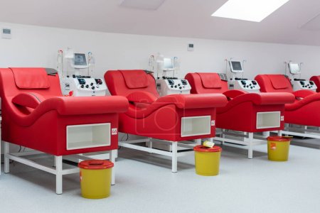 fila de sillas médicas modernas y cómodas cerca de máquinas de transfusión con pantallas táctiles, soportes de goteo con bolsas de infusión, cubos de basura en ambiente estéril de centro de donación de sangre