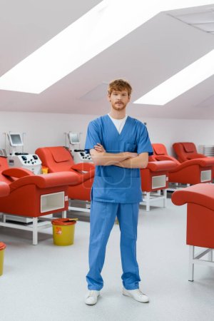 longitud completa de pelirroja y joven médico en uniforme azul de pie con los brazos cruzados cerca de cómodas sillas médicas, máquinas de transfusión automatizada y cubo de basura en el centro de donación de sangre
