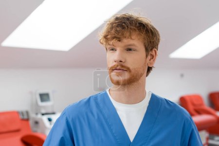 médico joven, pelirrojo y barbudo en uniforme azul de pie y mirando hacia otro lado cerca de las sillas médicas borrosas en el ambiente estéril de la estación de transfusión de sangre