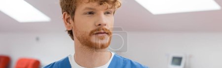 young, redhead healthcare worker in blue uniform looking away in sterile environment of laboratory in blood donation center on blurred background, banner