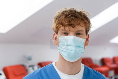 Photo for Young and redhead doctor with positive eyes expression, wearing medical mask and looking at camera near comfortable chairs on blurred background in blood donation center - Royalty Free Image