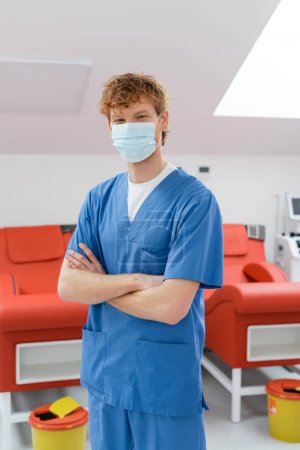 jeune médecin rousse en masque médical et uniforme bleu debout avec les bras croisés et regardant la caméra près des chaises médicales avec un design ergonomique et des seaux à ordures dans le centre de don de sang