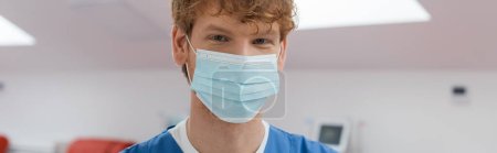 Photo for Young and redhead doctor with cheerful eyes expression, wearing medical mask and looking at camera in sterile environment of blood donation center on blurred background, banner - Royalty Free Image