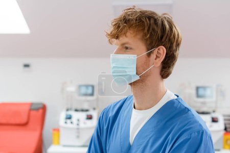 profil de rousse, jeune et positif médecin en masque médical et uniforme bleu regardant loin dans le centre de don de sang près des machines de transfusion avec moniteurs sur fond flou