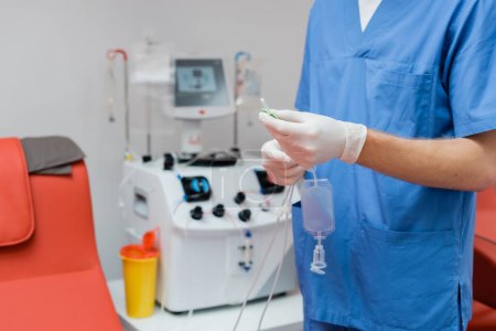 vista recortada del médico en uniforme azul y guantes de látex estériles que sostienen el conjunto de transfusión de sangre cerca de equipo automatizado borroso y taza de plástico en el laboratorio