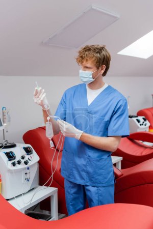 Rotschopf-Arzt in blauer Uniform, medizinischer Maske und Latexhandschuhen mit Transfusionsset in der Nähe von automatisierten Geräten und bequemen medizinischen Stühlen im Blutspendezentrum