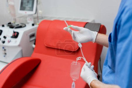 Teilansicht des Arztes in sterilen Latexhandschuhen mit Bluttransfusionsset in der Nähe des Medizinstuhls mit komfortablem ergonomischem Design und moderner Ausstattung auf unscharfem Hintergrund im Labor