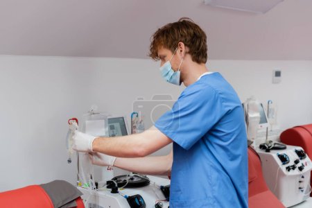 rothaarige Gesundheitshelferin in medizinischer Maske, blauer Uniform und Latexhandschuhen mit Transfusionsset in der Nähe von Tropfstühlen, medizinischen Stühlen und automatisierten Laborgeräten