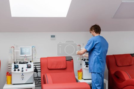 vista posterior del joven médico pelirrojo en uniforme azul cerca de máquinas de transfusión automatizadas, vasos de plástico, soportes de goteo y cómodas sillas médicas en el centro de donación de sangre