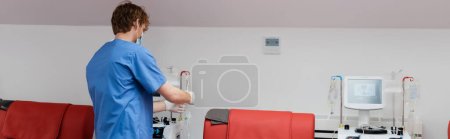 back view of redhead doctor in blue uniform, medical mask and latex gloves working with blood transfusion equipment near red medical chairs and drip stands in medical laboratory, banner