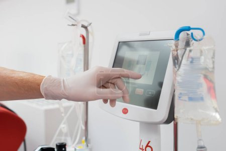 partial view of healthcare worker in latex glove operating modern automated transfusion machine with touchscreen near drip stands and infusion bags in medical laboratory
