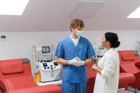 femme multiraciale et médecin rousse en uniforme bleu, masque médical et gants en latex parlant près de chaises médicales et machine de transfusion automatique dans le centre de don de sang