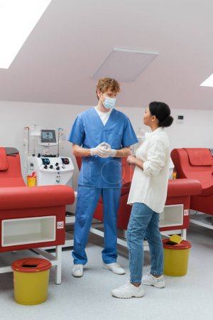 multiracial woman talking to redhead doctor in blue uniform, medical mask and latex gloves near red ergonomic chairs and automated transfusion machine in laboratory