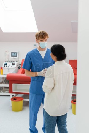 redhead doctor in medical mask, blue uniform and latex gloves talking to multiracial near medical chairs and automated transfusion machine on blurred background