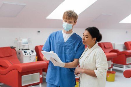 Junge rothaarige Ärztin in blauer Uniform, medizinischer Maske und Latex-Handschuhen zeigt der multirassischen Frau in der Nähe verschwommener Medizinstühle im Blutspendezentrum ein digitales Tablet