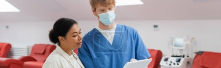 Multirassische Frau und rothaarige Ärztin in blauer Uniform und medizinischer Maske mit Blick auf digitales Tablet in der Nähe von Medizinstühlen und Bluttransfusionsgerät in der Klinik, Banner