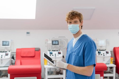 trabajador sanitario pelirrojo en uniforme azul, máscara médica y guantes de látex mirando a la cámara cerca de sillas médicas y máquinas de transfusión sobre fondo borroso en el centro de donación de sangre