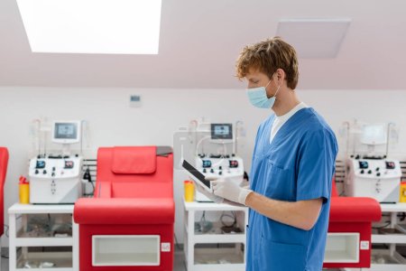 rothaarige Ärztin in medizinischer Maske, blauer Uniform und Latex-Handschuhen mit digitalem Tablet mit leerem Bildschirm in der Nähe von verschwommenen Medizinstühlen und automatischen Transfusionsmaschinen im Blutspendezentrum