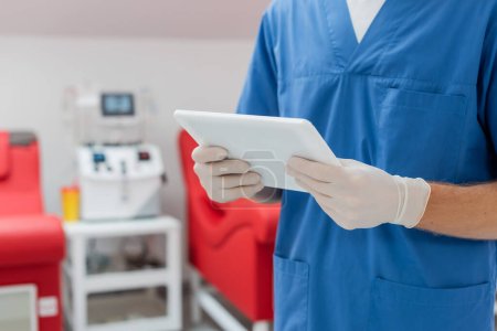 Ausgeschnittene Ansicht eines Arztes in blauer Uniform und Latexhandschuhen, der ein digitales Tablet in der Nähe verschwommener medizinischer Stühle und eines automatischen Transfusionsautomaten im Blutspendezentrum hält