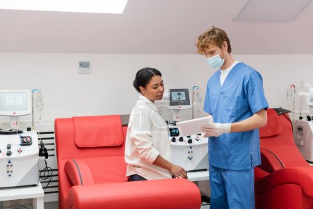 Arzt in medizinischer Maske, blauer Uniform und Latex-Handschuhen zeigt der multiethnischen Frau, die auf einem bequemen ergonomischen Stuhl in der Nähe von Transfusionsgeräten im Blutspendezentrum sitzt, ein digitales Tablet