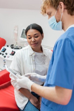 rothaarige Ärztin in medizinischer Maske, blauer Uniform und Latex-Handschuhen zeigt einer multirassischen Frau in der Nähe eines verschwommenen Transfusionsgeräts im Blutspendezentrum ein digitales Tablet 