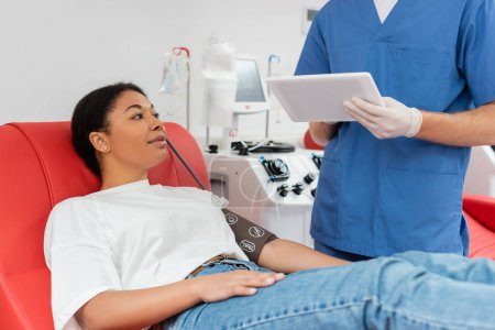 Arzt in blauer Uniform und Latexhandschuhen mit digitalem Tablet in der Nähe einer multiethnischen Frau in Blutdruckmanschette, die auf einem medizinischen Stuhl in einem modernen Labor sitzt