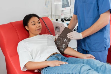 Gesundheitshelfer in blauer Uniform und Latexhandschuhen legt Blutdruckmanschette auf den Arm einer multiethnischen Frau, die auf einem bequemen medizinischen Stuhl in der Nähe eines automatischen Transfusionsautomaten im Krankenhaus sitzt 