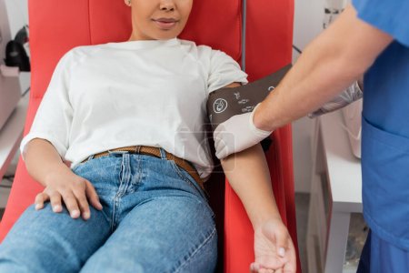 Teilansicht des Arztes in Latexhandschuhen, der die Druckmanschette auf einer multirassischen Frau einstellt, die auf einem bequemen medizinischen Stuhl in einer Bluttransfusionsstation sitzt, Patientenversorgung