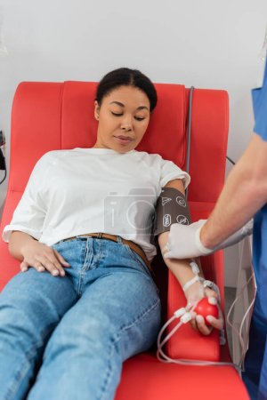 healthcare worker connecting blood transfusion set to multiracial woman sitting on comfortable medical chair in blood pressure cuff and squeezing rubber ball