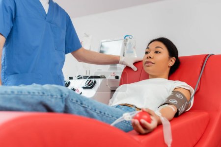 médico en uniforme azul de pie cerca de la mujer multirracial sentado en la silla médica cómoda cerca de la máquina de transfusión mientras dona sangre en el laboratorio médico