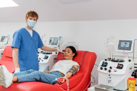 Arzt in medizinischer Maske, blauer Uniform und Latex-Handschuhen blickt in die Kamera in der Nähe von Transfusionsmaschinen und multiethnische Frau sitzt auf dem Behandlungsstuhl, während sie in der Klinik Blut spendet
