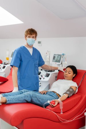 rothaarige Ärztin in medizinischer Maske und blauer Uniform blickt in die Kamera in der Nähe automatisierter Transfusionsmaschinen und multiethnischer Menschen, die auf dem Behandlungsstuhl sitzen und im Labor Blut spenden