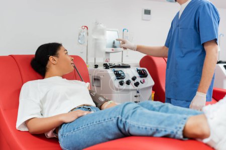 Gesundheitshelfer in blauer Uniform und Latexhandschuhen bedienen automatische Transfusionsmaschine in der Nähe einer Frau, die in einem medizinischen Labor Blut spendet