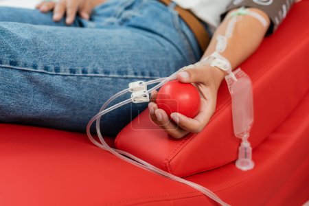 vista parcial de la mujer multirracial con transfusión conjunto celebración de bola de goma mientras está sentado en la silla médica cómoda durante la donación de sangre en el laboratorio