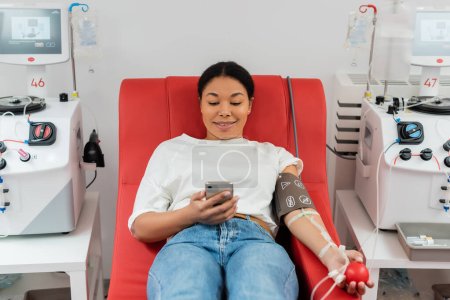 lächelnde multirassische Frau in Bluttransfusionsset mit Gummiball und Internet-Surfen auf dem Handy, während sie auf einem medizinischen Stuhl in der Nähe automatisierter Geräte im Labor sitzt